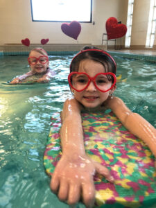 Picture of 2 swimmers on kickboards in the water together one in front of the other, its I heart Swimming week, so they have red headbands with hearts coming off the top of it and red heart shaped glasses on.