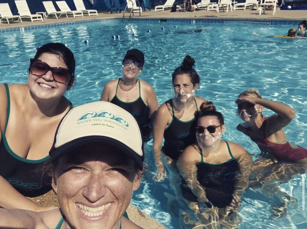 Picture of 6 Swim instructors smiling at the camera at the outdoor pool. It is very sunny out so some of the ladies are wearing sunglasses or hats, 
