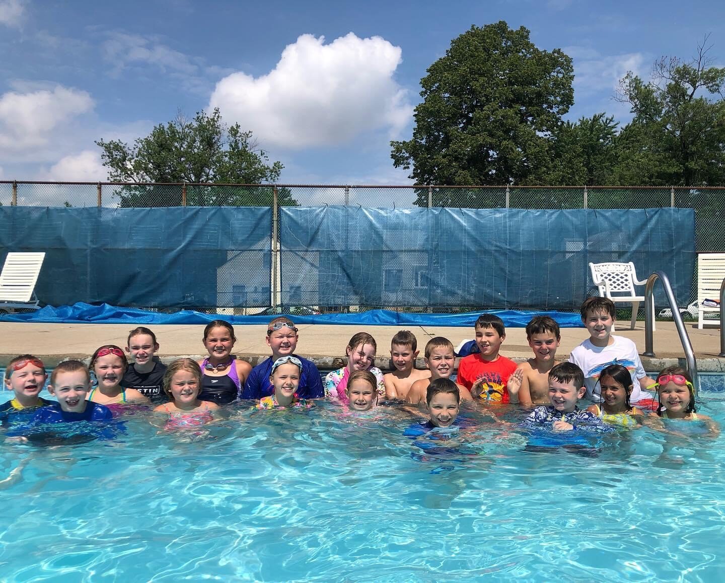 Picture of 19 Jr. Swim Team boys and girls in the water after practice. they are all smiling at the camera and it is sunny outside. They are the outdoor pool in evergreen park.